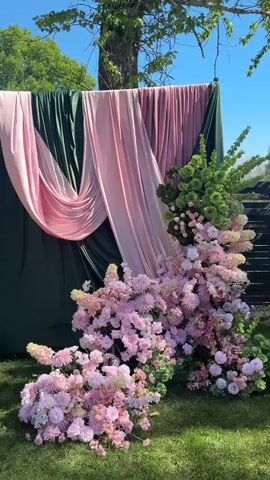 The video shows an outdoor setting with a decorative setup consisting of draped fabrics and floral arrangements. The backdrop is made of two colors of fabric: a dark green velvet and a lighter pink fabric that is draped in a flowing manner. The fabric backdrop is positioned against a backdrop of trees and a clear blue sky. In front of the fabric backdrop, a large floral arrangement is placed. The arrangement features various pink and white flowers, including what appears to be chrysanthemums and hydrangeas, as well as vibrant green foliage. The flowers are arranged in a cascading, layered pattern. The setting suggests the preparations for an event such as a wedding or photoshoot, and the floral arrangement and fabric drape creates an aesthetically pleasing setting.