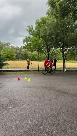 A cyclist, wearing red, maneuvers around orange and yellow cones in a paved area, with a group of observers standing in the background. The cyclist is riding a black bicycle. Behind them, a grassy field leads to trees under a cloudy sky. The observers include a person wearing a yellow jacket, someone in a red raincoat, and two other individuals, all standing on the far side of a low concrete wall. A few more trees are present in the scene, along with some bushes on the left side of the frame.