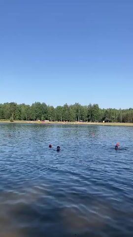 This video shows a sunny day at a lake in Hantsavichy, Belarus. The clear blue sky and lush green trees bordering the lake suggest warm weather, likely during the summer season. Several people are enjoying the water, some swimming, others wading near the shore. There are orange buoys in the water, marking swimming areas.

In the foreground, the water is calm with gentle ripples. Further out, groups of people are visible. Some individuals are playing with inflatable toys, including what appears to be a pink flamingo float. Children are also seen in the water, with one boy interacting with an adult and a ball. On the sandy shore, more people are relaxing and sunbathing. The scene conveys a sense of leisure and recreation, with individuals of various ages enjoying a day outdoors. The lighting indicates it is daytime, likely mid-morning to early afternoon, given the brightness and shadows. There is no visible text in the video.