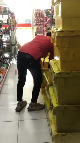 A person wearing a red shirt and black pants is bending over in a store aisle, reaching into a stack of yellow storage containers. Shelves stocked with various products are visible on the left side of the aisle. The floor is tiled, and the lighting suggests an indoor environment.