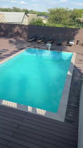A man in white shorts dives into a turquoise swimming pool. He is captured mid-air, a moment before he hits the water. The pool is surrounded by a wooden deck with several lounge chairs lined up along the back. Beyond the pool and deck, a brown wooden fence separates the area from a residential neighborhood with houses and trees under a bright blue sky with scattered clouds.