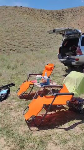 The video shows a campsite setting on a sunny day. In the foreground, two orange lounge chairs are set up next to a small folding table with drinks and other items. A silver SUV with its trunk open is parked behind the chairs. A green towel is draped over one of the chairs. Further back, a grill with food cooking over coals is surrounded by rocks. A blue plastic bag and some white packaging are visible on the grass near the grill. The environment appears to be a dry, grassy hillside. There are no people visible in the video.