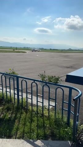 The video shows a small airport on a sunny day. In the background, a small airplane is moving on the runway, with mountains visible in the distance. A blue metal fence with planters lines the foreground, with grass and weeds growing around it. The sky is blue with scattered clouds.