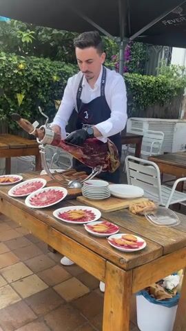 A man wearing a white shirt and a black apron with a logo is standing at a wooden table, slicing a large ham with a knife. The ham is secured in a metal stand. Several plates with thinly sliced meat and breadsticks are arranged on the table in front of the man. The background shows greenery and outdoor seating with white chairs. The man is wearing black gloves and is focused on his task. The lighting suggests it is daytime.