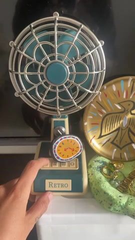 A hand with painted fingernails is shown pressing a button on a retro-style desk fan. The fan is teal and cream-colored, with a metal grille. A small, round rock painted with a clock face sits on top of the fan's base, near the button. To the right of the fan, a gold decorative plate with a bird design is visible. Below the plate, a pile of assorted jewelry, including rings and a watch, rests on a green, textured surface. The background appears to be a dark surface, possibly a television screen. The overall scene suggests a close-up shot of decorative items arranged together.