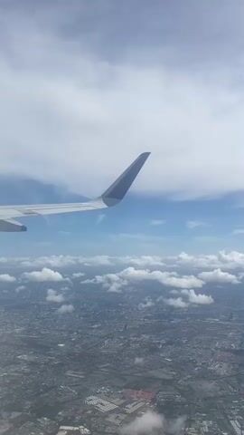The view from an airplane window shows the wing of the plane against a backdrop of blue sky and scattered clouds. Below, a sprawling city is visible. The scene is shot from a high angle, conveying the perspective of being airborne.