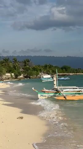 The video opens with a close-up view of waves gently lapping onto a sandy beach. A small outrigger boat, named "Mae Anon", is moored in the shallow turquoise water. The boat has a green hull and yellow outriggers, and a white canopy structure. The camera then pans slowly to the left, revealing a coastline dotted with palm trees and lush vegetation. In the background, distant mountains are visible under a partly cloudy sky. The beach itself is composed of light-colored sand. As the camera continues to pan, a few other boats can be seen further down the coast, and some buildings are nestled among the trees. The video concludes by panning further left, showing a lone person walking on the beach near a leaning palm tree and two white poles. The overall atmosphere is peaceful and tropical.