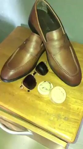 Here's a description of the video:

The video shows a pair of brown leather loafers, sunglasses, and two small, round wooden containers on a wooden surface. The sunglasses have gold-colored frames and dark lenses. One of the wooden containers is open, revealing a coiled, light green cloth inside. The other container has a label that reads "EVELYN & CRUSH". The surface appears to be part of a dresser or cabinet.