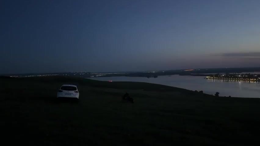 The scene is a wide shot of a dark, grassy hillside overlooking a wide river at dusk. The sky is dark blue, transitioning to a lighter color near the horizon. Along the riverbanks, lights from towns or cities are visible. A white car is parked on the left side of the hill. A person on a bicycle or motorcycle is visible in the center of the frame, also on the hillside. The landscape is mostly dark due to the low light.