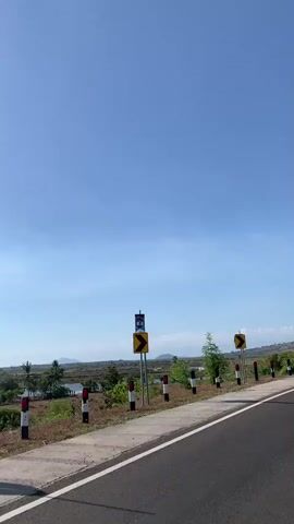 The video captures a view from a vehicle driving on a paved road. The sky is clear and blue. On the right side of the road, there are a few black and white striped posts, a yellow curved arrow sign, and a large telecommunications tower. Further down the road, there are trees and rolling hills in the background. A motorcycle is seen in the distance on the right side of the road. The vegetation along the roadside appears dry and sparse in some areas, with some green trees scattered throughout. There is a body of water visible on the left side of the road.
