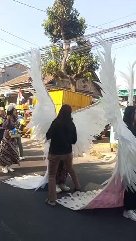 A group of people are walking down a street in what appears to be a parade or festival. In the foreground, a person wearing large white angel wings is being adjusted by another person. The person with the wings is wearing a pink shirt and dark pants, and the person adjusting them is wearing a black top and brown pants. Behind them, other people are walking, some carrying flowers. The background shows buildings and trees, with a clear blue sky above. The scene suggests a celebratory event.