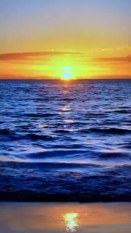 A sunset over a body of water. The sun is setting on the horizon, casting an orange and yellow glow across the sky and the water. The water is blue and choppy, with small waves. The reflection of the sun is visible on the water's surface. The scene is peaceful and serene.