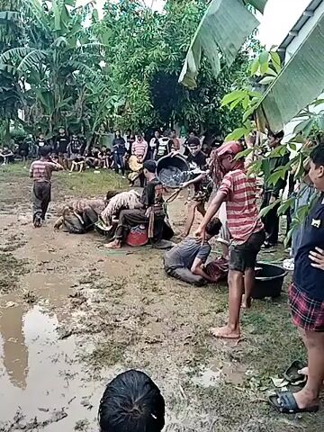 The video shows a group of people in a muddy area, possibly a field or yard. Some are sitting or kneeling in the mud, while others are standing around them. Many of the individuals are covered in mud. In the background, there is a crowd of onlookers standing and watching. There are trees and vegetation surrounding the area. One person is holding a long stick or whip, and another is carrying a bucket of water. The scene appears to be part of a ritual, a game, or some kind of organized activity.