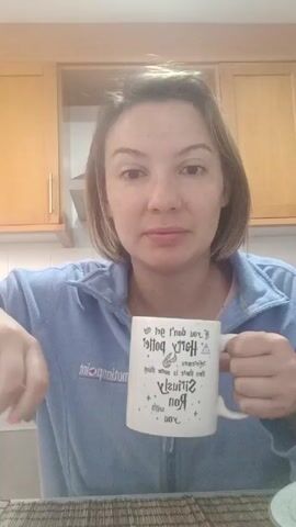 A woman with short, light brown hair and fair skin is sitting at a table with a woven placemat in front of wooden cabinets. She wears a light blue fleece jacket. The woman is holding a white mug with text on it, which she turns to face the camera. She says something to the camera and then takes a drink from the mug.
