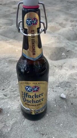A close-up, eye-level shot focuses on a bottle of Hacker-Pschorr Münchner Gold beer, standing upright in fine sand. The bottle has a dark amber glass body and a swing-top closure with a red ceramic stopper. A golden label adorns the bottle, featuring the Hacker-Pschorr logo and text in German. The background consists of more sand, with some small pebbles scattered around. In the first few seconds, a hand enters the frame from the left and touches the metal wire of the swing-top closure. The hand then moves to grip the bottle and begins to lift the swing-top mechanism. The video ends with the swing-top partially open, poised to release the beer.