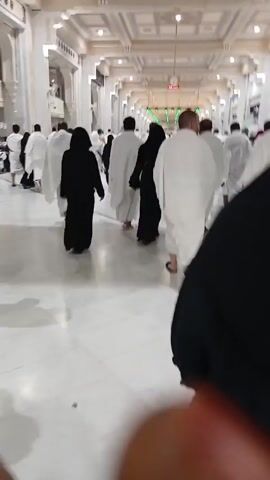 The video shows a large group of people walking inside a large, well-lit building. Most of the people are wearing white robes, while some are wearing black garments. The building appears to be a religious site, with a patterned ceiling and bright lights. The crowd is moving in the same direction, suggesting they are part of a procession or pilgrimage. The camera is handheld and moves along with the crowd, providing a first-person perspective.