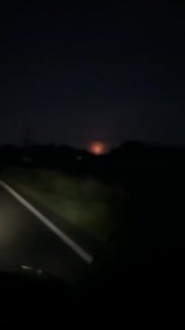The video captures a view from a vehicle at night, likely on a road. The headlights illuminate a white line on the asphalt in the foreground. The middle ground shows dark silhouettes of trees and vegetation. In the background, a bright, reddish-orange light is visible above the treeline, possibly indicating a distant light source, sunset, or moonrise. The sky is dark and overcast. The video is short, showing a slight movement of the camera, suggesting the vehicle is in motion.