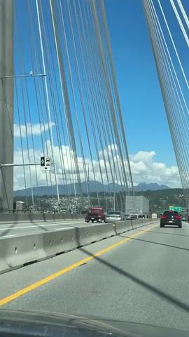 The video shows a highway scene from the perspective of a vehicle driving on a bridge. The bridge is supported by numerous cables that stretch upwards and to the right. The road is paved with asphalt and has a yellow line dividing the lanes. On the left side of the road, there is a concrete barrier. Cars and trucks are driving in the same direction as the viewer. In the distance, there are mountains covered in trees, and a clear blue sky with white clouds. The video appears to be shot on a sunny day.