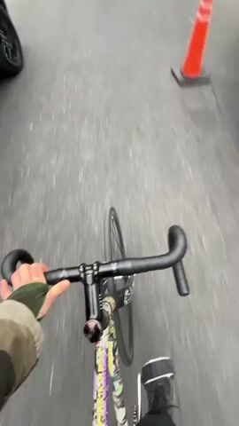 A first-person perspective video shows someone riding a bicycle on a paved street. The person is wearing a green jacket. To the left is a gray SUV. There are two people in bright yellow vests, and orange traffic cones. The cyclist proceeds past these people and cones. Ahead, another cyclist crosses the street via the crosswalk, moving past a parked truck and a tow truck with flashing lights. Overhead are tree branches against a gray sky.