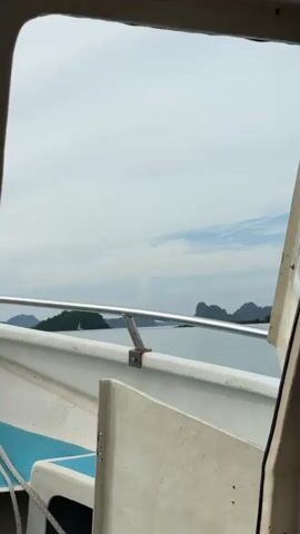 The video shows a view from inside a boat, looking out through an opening. The opening frames a body of water with several islands in the distance. The islands are covered in green vegetation and have rocky, irregular shapes. The water is a calm, blue-gray color, and the sky is mostly cloudy with some patches of blue. The boat itself is white with blue seats visible in the foreground. A metal railing runs along the side of the boat. The camera appears to be stationary, and the boat is moving, as indicated by the slight changes in the perspective of the islands. There are no people visible in the video.
