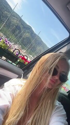 A young woman with long blonde hair and sunglasses is sitting in a car, looking at the camera. The car has a sunroof open, revealing a beautiful view of mountains, a harbor with sailboats, and a town with red-tiled roofs. There are planters filled with colorful flowers along a railing. The woman is wearing a white shirt. The scene appears to be a sunny day.