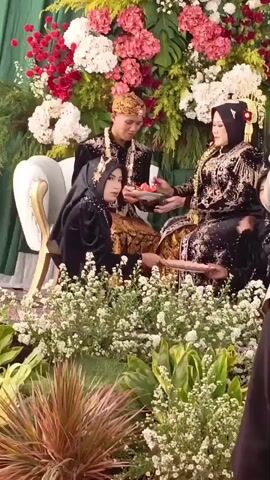 A wedding ceremony is depicted with a couple seated on chairs, adorned in traditional attire. The groom wears a golden headpiece and a black outfit with ornate detailing. The bride is seated next to him, also in traditional wedding attire, including a black veil with gold embroidery and a tall headdress. She is holding a plate with red fruits and feeding one to the groom. Another woman in a black veil and traditional clothing is seated at the groom's left, looking at the couple. The background is decorated with abundant floral arrangements, featuring red and white flowers, and lush greenery. The foreground is filled with small white flowers and other plants, creating a natural setting. The lighting appears to be bright, suggesting an indoor or covered outdoor event.