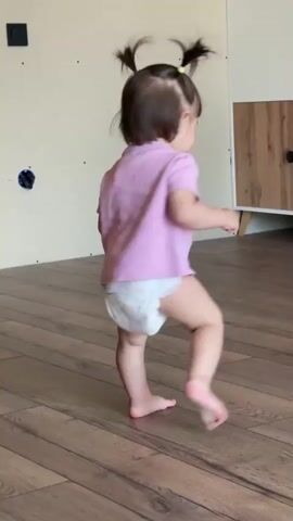 A toddler girl with brown hair in pigtails and a pink t-shirt with a graphic print and diaper is walking barefoot on a wood floor in a room undergoing renovation. The wall is unfinished drywall. She turns to smile briefly at the camera, then turns away to continue walking.