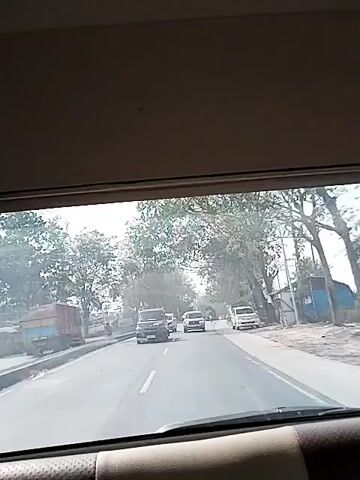 The video shows a view from inside a vehicle traveling on a two-lane road. Several cars and trucks are visible on the road, moving in the same direction as the camera vehicle. The road is flanked by trees and vegetation. On the right side of the road, there are buildings, including a blue structure and a yellow truck. A person on a red motorcycle is also seen on the right side. The lighting suggests it is daytime, possibly sunny. The camera is positioned to capture the road ahead through the windshield.