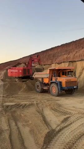 In a sand quarry, a large red Hitachi excavator is actively digging and loading sand into an orange dump truck. The excavator's arm is extended, its bucket filled with sand, poised to deposit its load into the truck's bed. The dump truck, identified with the number "28" on its front, is positioned to receive the sand. The setting is a dry, sandy environment with tall, layered sandbanks dominating the background. The sky above is a pale, clear blue, suggesting it is either early morning or late afternoon. The ground is uneven and covered in sand, with visible tire tracks from heavy machinery. The overall scene depicts an industrial operation in a quarry, with heavy equipment engaged in excavation and transportation. No people are visible in the frame. The time of day appears to be either dawn or dusk due to the lighting and sky conditions. The location is identified as Tatsinskaya, Russia.