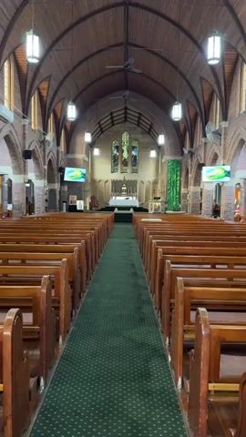 The video shows the interior of a church, looking from the back towards the altar. Wooden pews are arranged in rows on either side of a green carpeted aisle. The ceiling is arched and made of wood, with hanging lights and ceiling fans. Stained glass windows are visible at the far end of the church, above the altar. There are large screens mounted on the walls on either side of the aisle, displaying colorful images. The altar area is elevated and features a white cloth-covered table and other religious items.