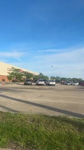 The video shows an outdoor scene in a large paved area, likely a parking lot, on a clear, sunny day. Several police SUVs are visible, some with flashing blue and red lights. In the initial frames, they appear to be moving slowly in a circular pattern or formation. One black police SUV has a distinctive striped design on its rear.

In the background, there's a large commercial building with light-colored walls and some brick accents. A sign with "AMC" is visible on the building, suggesting a movie theater. Other commercial signs are present, indicating a shopping center with "BURLINGTON," "TARGET," and "AMC CINEMAS" listed. Traffic cones are placed on the ground, and a "20" speed limit sign is visible. The area is surrounded by some trees and grassy patches. The overall impression is an organized presence of law enforcement, possibly for an event, training, or response at a commercial complex.