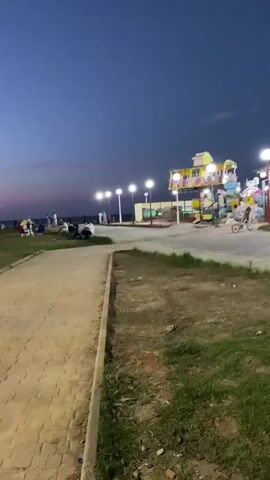 The video shows an amusement park scene at dusk. Several colorful amusement rides, including a roller coaster and a carousel, are visible. Lights are illuminated on the rides and along a walkway. People are scattered throughout the park, some on the rides, others walking or standing around. A person is riding a bicycle on a paved path. The ground is covered in grass and dirt, and there are a few scattered rocks. The sky is a deep blue, indicating the transition from day to night.