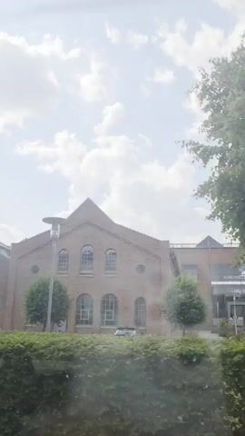The video shows a view from a vehicle moving past buildings. The buildings are made of brick and have a triangular roof. There are several windows on the buildings. There are trees and bushes in front of the buildings. The sky is cloudy. There are banners on the buildings with text. One banner says "ANJOCK" and another says "IT-Zentrum". There is a signpost in front of one of the buildings. The video is shaky, suggesting it was filmed from a moving vehicle.