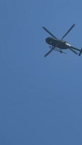 The video shows a dark-colored police helicopter flying in a clear blue sky. The helicopter is seen from below, with the word "POLICE" visible on its side. The helicopter's rotors are spinning as it flies overhead. The camera angle is low, looking upwards towards the helicopter.