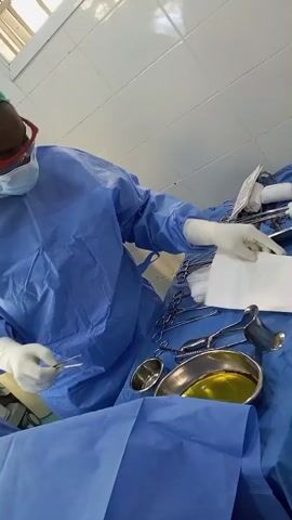 A person in a blue surgical gown, green surgical cap, mask, safety glasses, and white gloves is in a medical setting. They are holding a pair of surgical forceps in their left hand and a white paper in their right hand. The person is leaning over a surgical tray covered in blue drapes, which contains various surgical instruments and a metal bowl filled with a yellow liquid. The background appears to be a tiled wall. The person's actions suggest they are preparing or organizing surgical tools.