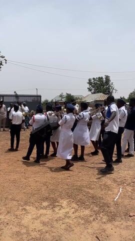 In a sunlit outdoor scene on an unpaved, reddish-brown dirt ground, a marching band is actively performing and moving from left to right. The band members, a mix of men and women, are dressed in white and blue uniforms, with some wearing white shirts and dark trousers. They play various brass instruments, including trumpets and trombones, as well as percussion instruments like a bass drum and a snare drum. The bass drum has "PREMIER" written on its side.

Several children, some in blue uniforms, are standing on the right, observing the band. Other adults are also visible in the background, either watching or accompanying the band. In the background, there's a black metal fence, some simple buildings, and sparse trees under a bright, clear sky. A dark-colored vehicle (possibly a van or SUV) is parked behind the onlookers on the right. The overall atmosphere suggests a community event in Laduran Foron, Nigeria.