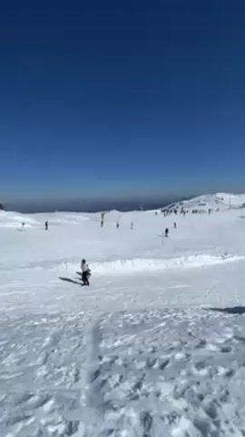 The video shows a ski slope on a sunny day. The sky is clear and blue. The ground is covered in snow, with many skiers and snowboarders visible in the distance. In the foreground, a woman and two children are standing on the snow, one of the children sitting on a sled. The camera pans slightly to the right. There are ski lifts and buildings visible in the background.