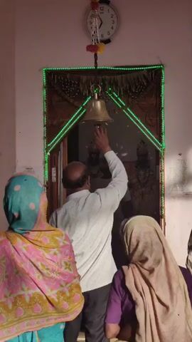 A man, facing away from the camera, rings a brass bell hanging from a chain. The bell is suspended in front of a doorway adorned with blue lights and decorative garlands. Inside the doorway, two figures, possibly statues, are visible. The man is wearing a white shirt. Three other people, partially visible and facing the doorway, are also present. One person to the left wears a pink and yellow patterned garment, and another person in the foreground to the right wears a beige head covering. A clock is mounted on the wall above the doorway.