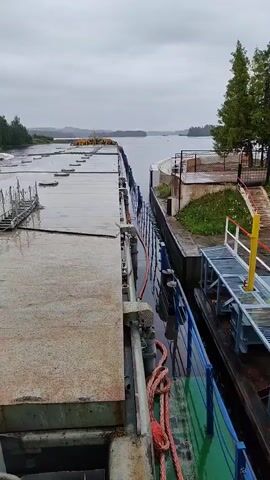 The video shows a view from the deck of a large barge looking towards the stern. The barge is alongside a concrete dock or quay, with another vessel visible to the right. The sky is overcast, and the ground is wet, suggesting it has rained. There are some metal structures on the barge and some railings on the dock. In the distance, there is a body of water with land on the other side.