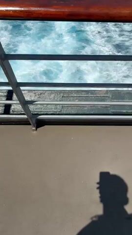 A video taken from a moving boat shows a wake of white foam trailing behind on the dark blue ocean. The camera angle is from behind a wooden railing, showing a portion of the boat's deck in the foreground. In the distance, the horizon is visible with a clear blue sky and some white clouds. There is a faint shadow of a person holding a phone in the bottom right corner of the first frame.
