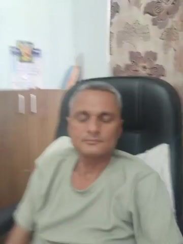 A man with graying hair and a light green t-shirt sits in a black office chair. He is looking at the camera. Behind him, there is a wooden cabinet with a calendar on it, a curtain with a floral pattern, and a white pillow on the chair. In one frame, the man raises his hand and makes a gesture with his fingers.