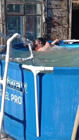 Two women are in a blue inflatable pool. One woman is wearing a black sports bra and purple shorts, and the other is wearing a pink and black sports bra and purple bottoms. They are embracing and appear to be playing or wrestling in the water. A third person, a woman with short hair, is visible at the edge of the pool, looking at the camera. The background shows a rustic wooden building and a brown fence. The sun is shining, creating bright lighting conditions.
