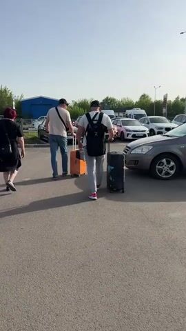 Three people are walking through a parking lot, pulling luggage. Two men are walking ahead with suitcases on wheels and a woman is walking behind them. The man in the front has a backpack. There are cars parked in the lot on either side of them, including a Kia and several BMWs. There is a blue building visible in the background. The sky is pale blue.