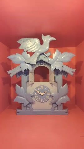 A person appears in the open doorway of a large, pale blue cuckoo clock. The clock is decorated with stylized leaves and has a carved bird perched on top. The background is a solid pink color, creating a vibrant and slightly surreal setting. The person is wearing sunglasses and appears to be looking out from the clock. The video seems to be a short, playful loop or a time-lapse, showing the person briefly appearing and disappearing from the clock's opening.