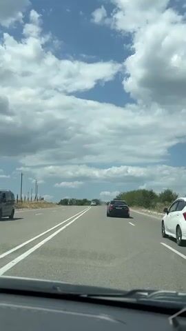 The video shows a view from inside a vehicle traveling on a paved road. The road has white double lines marking the lanes. In front of the vehicle, several other cars are visible, including a black sedan in the right lane and a white sedan further ahead in the left lane. The landscape on either side of the road is lined with green bushes and trees, with sparse vegetation and utility poles visible on the left. The sky is a bright blue with scattered white clouds. The overall environment appears to be a rural or semi-rural area on a clear, possibly warm day. The video captures the forward motion of the vehicle, indicating it is in transit.