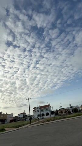 The video presents a static, low-angle shot of a street in a residential area on a day with a sky full of puffy, white altocumulus clouds against a backdrop of light blue. The street is asphalt and features a low, grassy verge separating it from the houses. Power lines stretch across the sky, supported by wooden poles. The houses vary in style, mostly one or two stories, with a mix of white and earth-toned facades. Some cars are parked in front of the houses. The sun appears to be setting or rising, as it casts a subtle warm glow through the clouds. The overall atmosphere is quiet and peaceful.