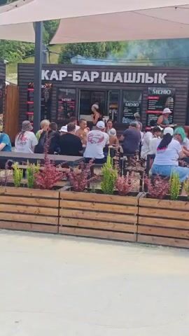 A group of people are gathered at an outdoor food court with several kiosks. The kiosks have signs in Russian, indicating they serve shashlik, burgers, waffles, blini, and coffee. Some people are sitting at tables under a canopy, while others are walking through the area. In the foreground, two individuals push a stroller, and a man walks by holding a bottle. The environment appears to be a recreational area, possibly near a beach or park, with trees visible in the background.