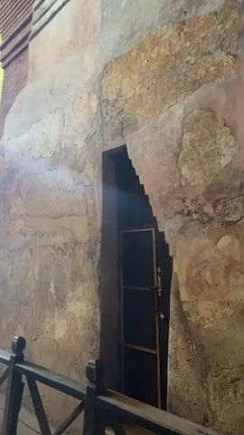 The video shows a close-up of a weathered wall inside what appears to be an old structure. The wall has faded frescoes featuring repetitive circular patterns.  A dark doorway with a metal security gate is visible in the background, letting in a stream of light. A wooden railing runs along the bottom of the frame, suggesting the area is preserved or restricted. The overall impression is of an ancient, possibly historical, site. There are no people visible in the video.