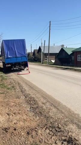 The video shows a street in a rural village under a clear blue sky. A blue road sign indicates directions and distances to Suksun and Syzganka. Houses of varying materials and states of repair line the street, with overhead power lines running along the road. The road itself is paved but shows signs of wear and tear. Vegetation grows on the roadside. The camera moves along the road, showcasing the village houses. A motorcycle is seen at a distance on the road.