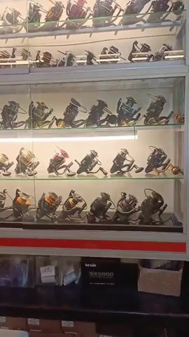 The video shows a display case filled with fishing reels. The reels are arranged on multiple shelves within the case. The shelves are illuminated, and the reels are shiny, suggesting they are new or well-maintained. The display case is made of glass, allowing a clear view of the merchandise. Below the display case, there are boxes and other fishing-related items. The overall impression is that of a fishing equipment store.